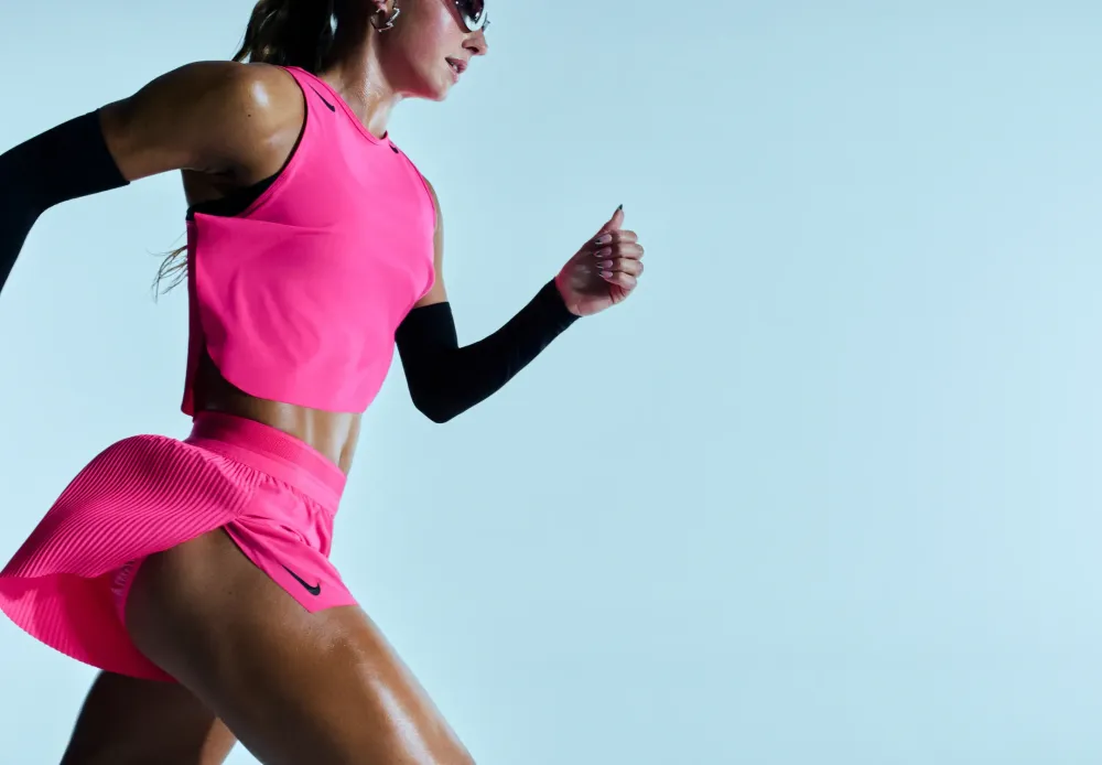 Runner in a pink Nike crop top and pleated shorts sprinting against a pale blue backdrop